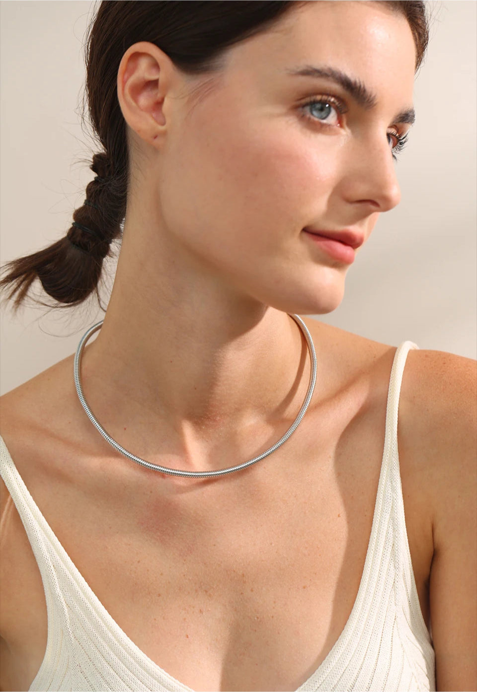 A close-up shot of a woman's neck showcasing a minimalist silver choker necklace with a smooth, polished finish. The elegant jewelry piece sits perfectly around her collarbone, reflecting light beautifully for a sleek and modern look.