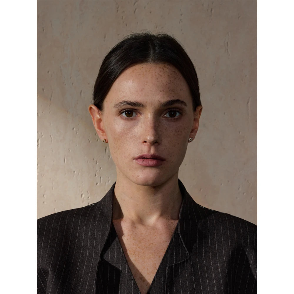 A refined pair of gold stud earrings with shimmering gemstones accentuates the woman’s natural beauty. Styled with a dark pinstriped blazer, these earrings offer a perfect balance of modern elegance and timeless charm.