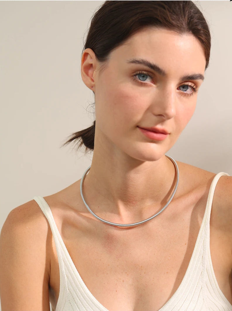 A close-up of a woman's neck and collarbone area, highlighting a sleek silver choker necklace. The necklace has a smooth, polished finish, sitting elegantly around her neck. Its minimalist and modern design makes it a versatile jewelry piece, perfect for both casual and formal wear. The soft lighting enhances the reflective quality of the silver, drawing attention to the elegant craftsmanship.
